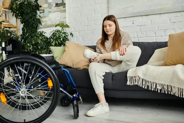 A young woman in a wheelchair puts on her sneakers while sitting on a couch in her living room. — Stock Photo