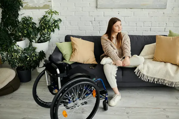 A young woman sits on a couch in her living room, her wheelchair nearby. — Stock Photo