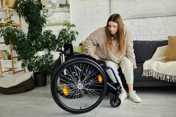 Eine junge Frau im Rollstuhl sitzt in ihrem Wohnzimmer und blickt auf ihren Rollstuhl. — Stockfoto