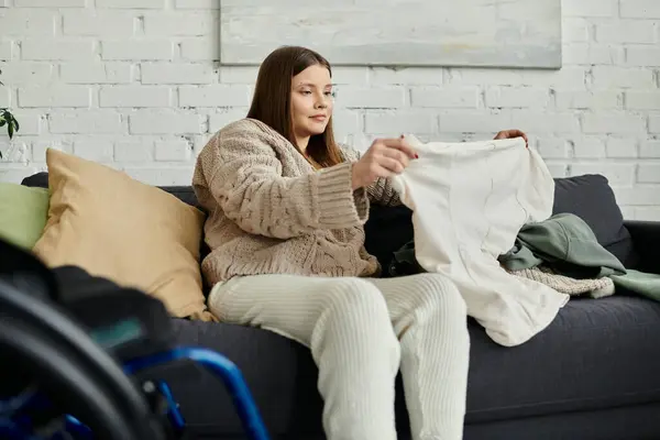 A young woman in a wheelchair sits on a couch in her living room, folding laundry and enjoying a moment of quiet. — Stock Photo