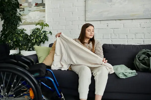 Une jeune femme est assise dans son fauteuil roulant dans son salon, pliant une couverture blanche. — Photo de stock