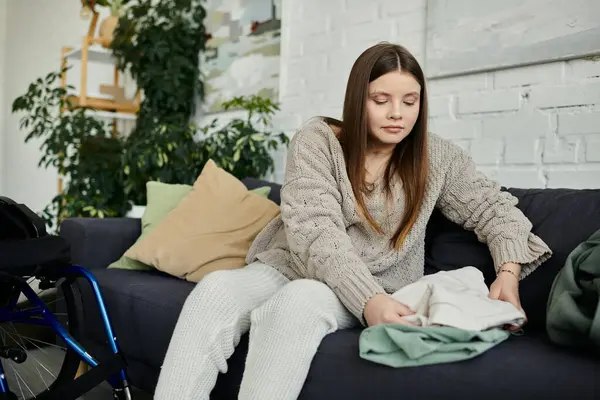 A young woman sits on a couch in her living room, folding laundry in her wheelchair. — Stock Photo