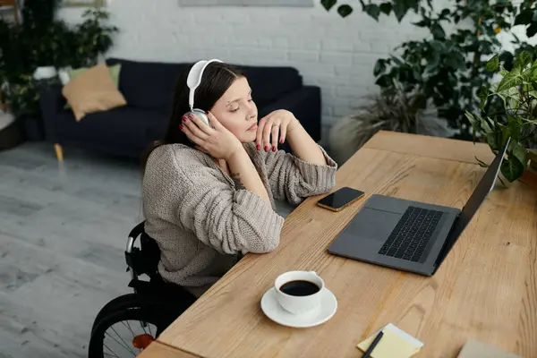 A young woman in a wheelchair sits at a desk, listening to music with headphones, enjoying a coffee break. — Stock Photo