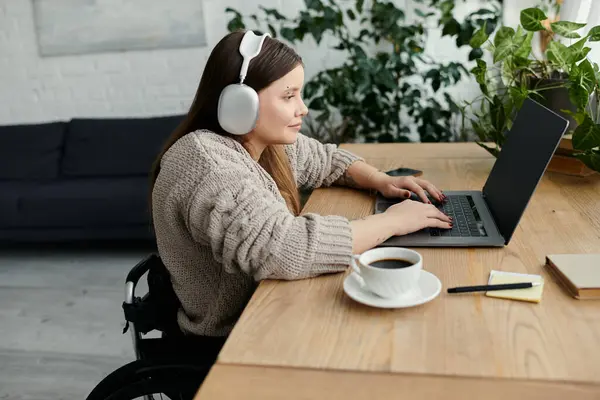 Una giovane donna su una sedia a rotelle lavora sul suo computer portatile mentre indossa cuffie a casa. — Foto stock