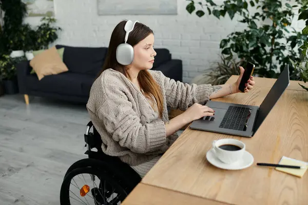 A young woman sits in a wheelchair at a desk in her home, working on her laptop. She is wearing headphones and looking at her phone. — Stock Photo