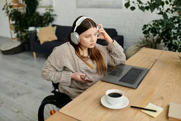 Eine junge Frau im Rollstuhl sitzt an ihrem Schreibtisch, trägt Kopfhörer und blickt nachdenklich auf ihren Laptop.. — Stockfoto