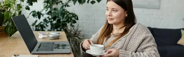 Eine junge Frau im Rollstuhl genießt zu Hause eine Tasse Kaffee. Ein Laptop liegt offen auf dem Tisch vor ihr. — Stockfoto