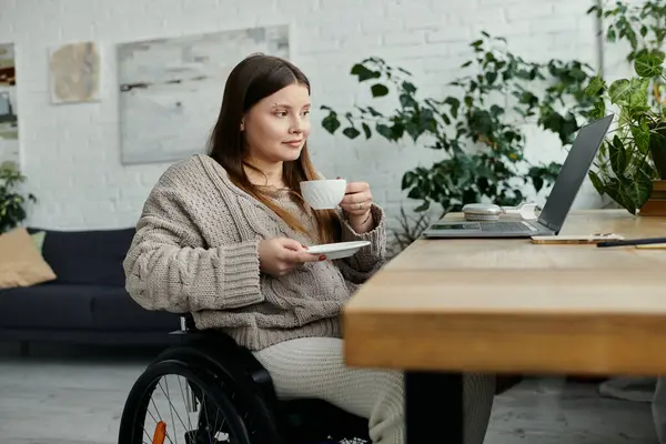Eine junge Frau im Rollstuhl genießt eine Tasse Kaffee am heimischen Schreibtisch. — Stockfoto