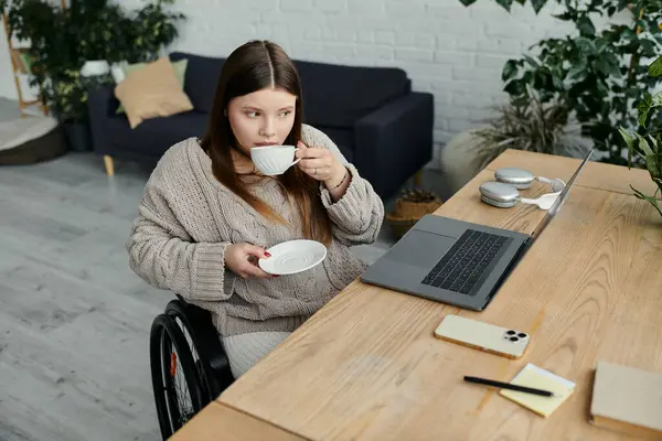 A young woman in a wheelchair enjoys a cup of coffee at her desk at home. — Stock Photo