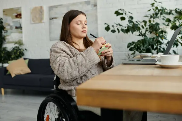 A young woman sits in a wheelchair at home, writing notes on a notepad. — Stock Photo