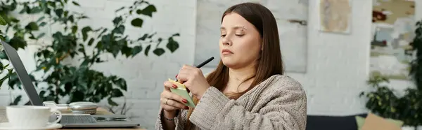 Una joven en silla de ruedas se sienta en una mesa escribiendo notas con un bolígrafo y un pequeño bloc de notas. — Stock Photo