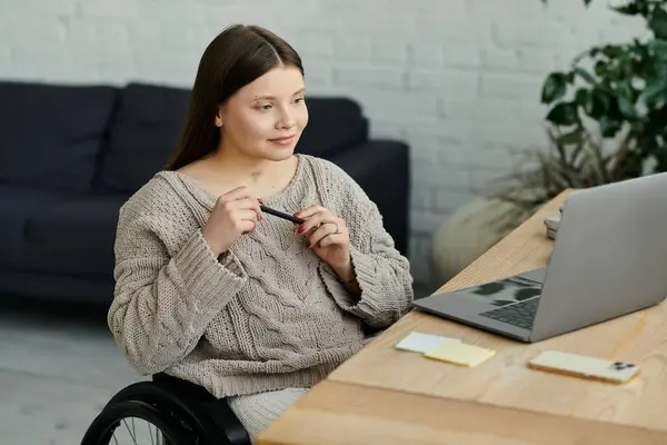 Eine junge Frau im Rollstuhl sitzt zu Hause an ihrem Schreibtisch und konzentriert sich mit einem Stift in der Hand auf ihre Arbeit. — Stockfoto