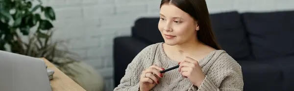 Una giovane donna si siede a un tavolo nella sua casa, utilizzando un computer portatile, tenendo la penna — Foto stock