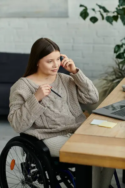 Una giovane donna si siede sulla sedia a rotelle, lavorando ad una scrivania. Indossa un maglione e usa un computer portatile e una penna. — Foto stock