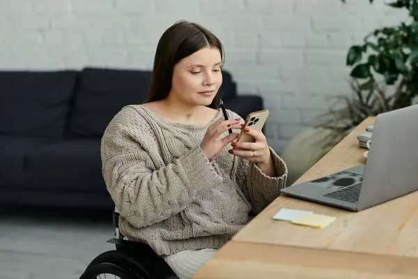 A young woman in a wheelchair sits at a table in her home, using her smartphone. — Stock Photo