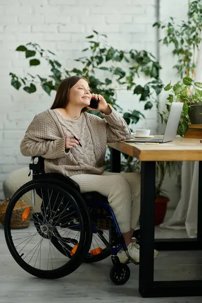 A young woman in a wheelchair sits at a table, smiling while talking on her phone. She is at home in a comfortable and bright setting. — Stock Photo