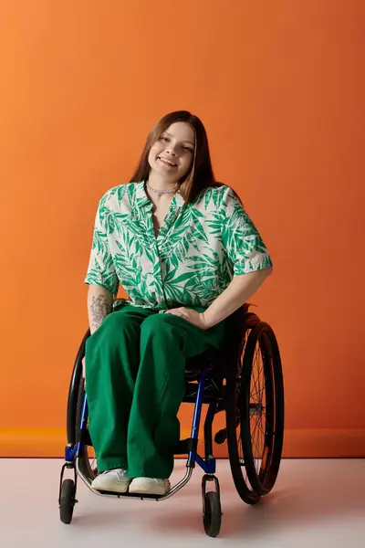 Une jeune femme dans une tenue verte vibrante s'assoit en toute confiance dans un fauteuil roulant, souriant à la caméra sur un fond orange vif. — Photo de stock