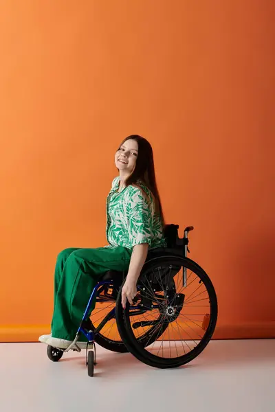 Eine junge Frau mit langen Haaren sitzt im Rollstuhl vor orangefarbenem Hintergrund, grün gekleidet und lächelt selbstbewusst. — Stockfoto