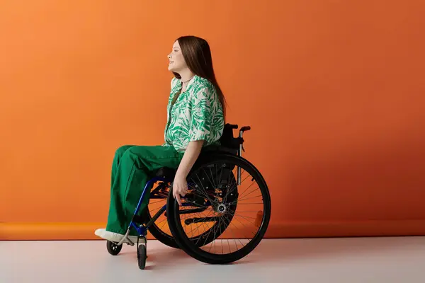 Молодая женщина в ярко-зеленом наряде уверенно сидит в инвалидном кресле на ярко-оранжевом фоне. — стоковое фото