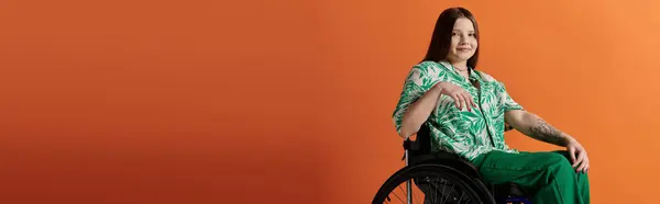 Una giovane donna, vestita con una vibrante camicia verde e pantaloni, siede fiduciosa su una sedia a rotelle su uno sfondo arancione. — Foto stock