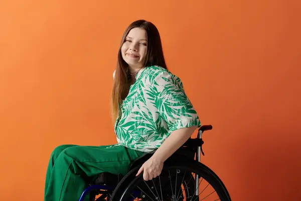 Eine behinderte junge Frau sitzt anmutig in einem leuchtend grünen Kleid vor orangefarbenem Hintergrund und strahlt Schönheit und Stärke aus.. — Stockfoto
