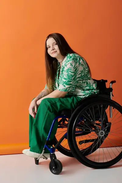 Eine selbstbewusste junge Frau mit langen braunen Haaren, grünem Hemd und grüner Hose sitzt im Rollstuhl vor orangefarbenem Hintergrund. — Stockfoto