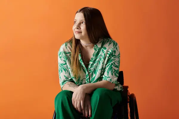 Una joven con el pelo castaño largo y una camisa verde vibrante se sienta en una silla de ruedas sobre un fondo naranja, sonriendo y mirando hacia arriba. — Stock Photo