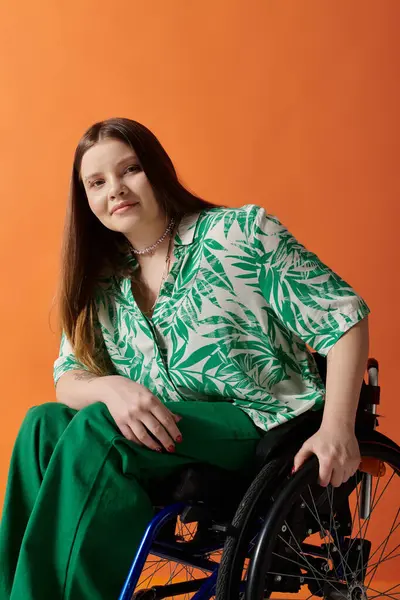 A young woman wearing green clothing with a leafy pattern smiles confidently while sitting in a wheelchair against an orange background. — Stock Photo