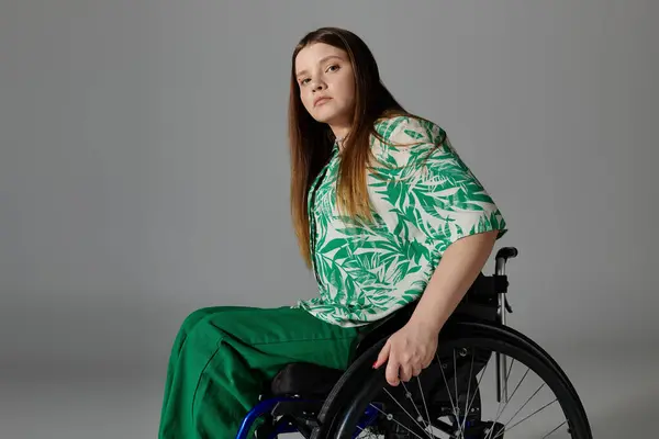 Une jeune femme vêtue d'une chemise verte et d'un pantalon s'assoit dans un fauteuil roulant sur fond gris. — Photo de stock