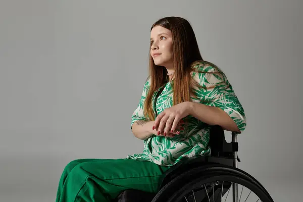 Young woman in green shirt in wheelchair gazes thoughtfully to the left against grey background. — Stock Photo