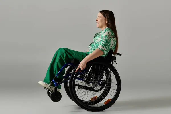 Una giovane donna vestita di verde siede su una sedia a rotelle su uno sfondo grigio, sorridente. — Foto stock