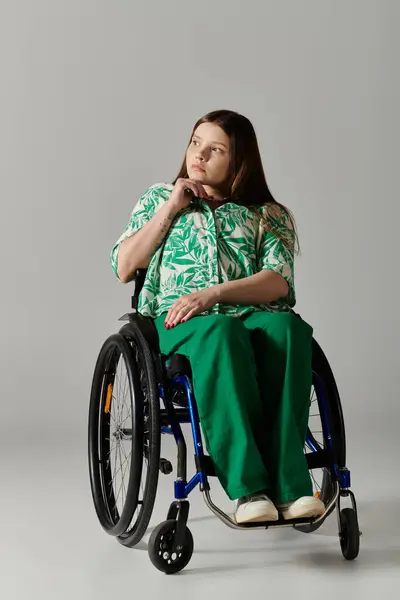 A young woman wearing a green patterned shirt and green pants sits in a wheelchair, looking thoughtfully towards the side. — Stock Photo