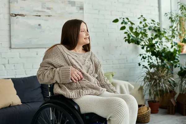 Une jeune femme en fauteuil roulant est assise dans un salon, portant un pull confortable et regardant par la fenêtre. — Photo de stock