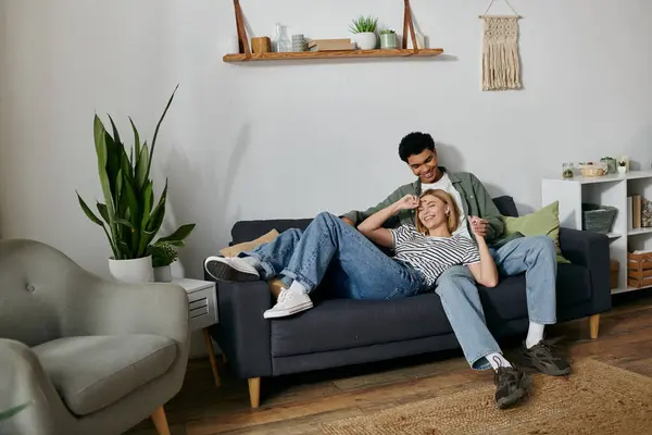 A young multicultural couple laughs and enjoys each others company on a comfortable couch in their modern apartment. — Stock Photo