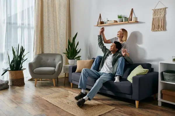 A young multicultural couple celebrates with raised arms, sitting on a sofa in a modern apartment. — Stock Photo