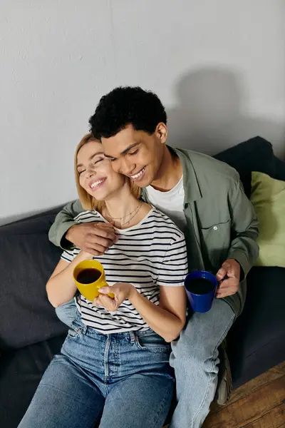 A young interracial couple relaxes on a couch in a modern apartment, sharing coffee and a cozy moment together. — Stock Photo