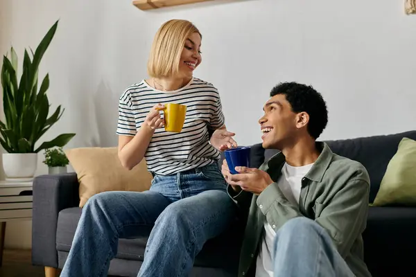 A young multicultural couple sits on a couch in their modern apartment, enjoying coffee and conversation. — Stock Photo