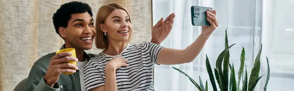 A young, multicultural couple enjoys a cozy moment in their modern apartment, taking a selfie with big smiles. — Stock Photo