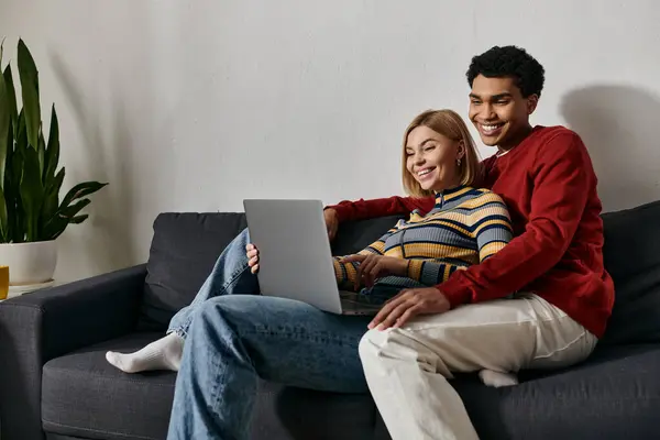 Un couple multiculturel heureux sourit tout en se relaxant ensemble sur un canapé et en naviguant sur Internet sur un ordinateur portable. — Photo de stock