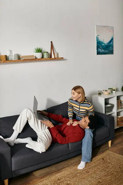 A happy multicultural couple relaxes on a couch in a modern apartment, the woman sitting on the male lap as he uses a laptop. — Stock Photo