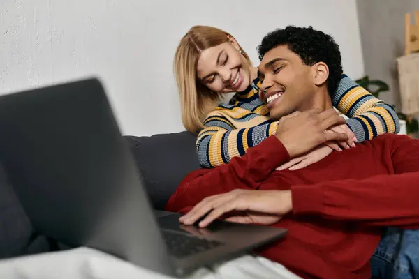 A happy, multicultural couple cuddles and laughs together while working on a laptop in their modern apartment. — Stock Photo