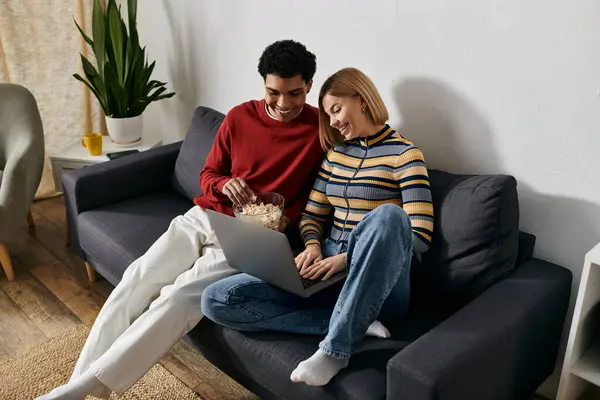 A happy multicultural couple relaxes on a couch in a modern apartment, enjoying a movie and popcorn. — Stock Photo