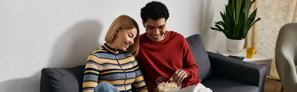 A multicultural couple relaxes on a couch in their modern apartment, enjoying a bowl of popcorn together. — Stock Photo