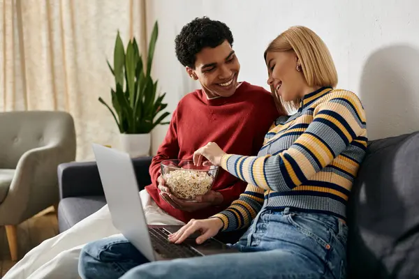 A happy multicultural couple enjoys a movie night in their modern apartment, sharing a bowl of popcorn and a laptop. — Stock Photo