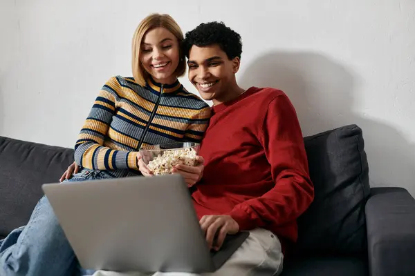 Um casal feliz e multicultural desfruta de uma noite aconchegante juntos, assistindo a algo em seu laptop e compartilhando uma tigela de pipocas. — Fotografia de Stock