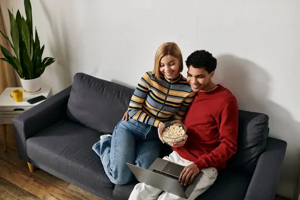 Una feliz pareja multicultural se acurruca en un sofá en un apartamento moderno, disfrutando de una noche de cine junto con palomitas de maíz y un ordenador portátil. - foto de stock