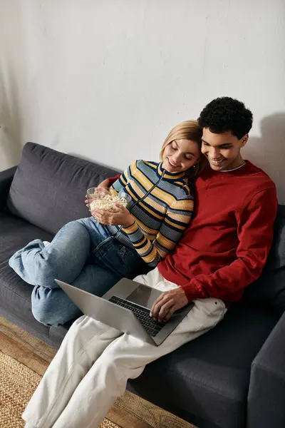 A happy multicultural couple enjoys quality time together on a modern couch, sharing popcorn and browsing on a laptop. — Stock Photo