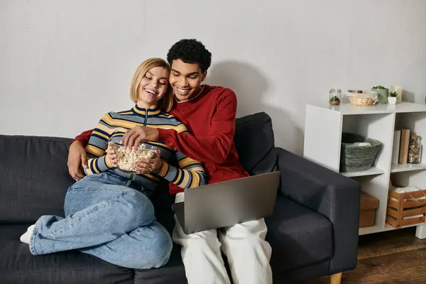 A multicultural couple enjoys a movie night at home, sharing popcorn and laughter on a comfortable couch. — Stock Photo