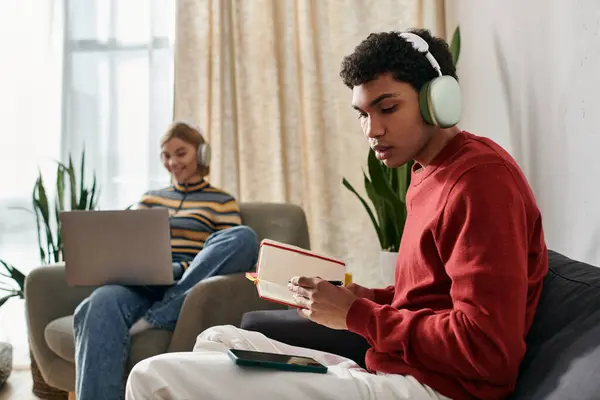 Una pareja multicultural se relaja en su apartamento moderno. Él usa auriculares y escribe en un cuaderno, ella trabaja en su computadora portátil. - foto de stock