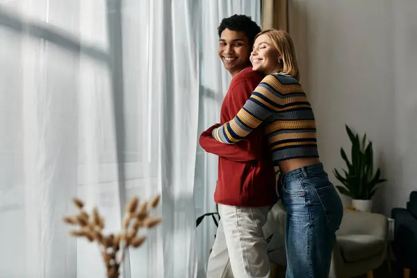 A happy, multicultural couple embraces by a window in their modern apartment. — Stock Photo